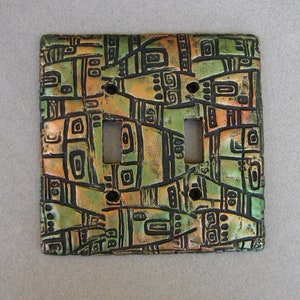 May include: A ceramic light switch cover with a geometric pattern in shades of green, brown, and orange. The pattern is reminiscent of ancient art.
