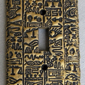 May include: A gold light switch cover with a black and gold design of hieroglyphics. The switch cover is made of ceramic and has a textured surface.