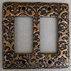 May include: A double switch plate with a decorative gold and black damask pattern. The plate has a raised, textured design.