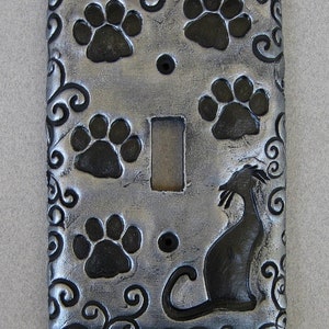May include: A silver light switch cover with a black cat silhouette and paw prints. The cover has a decorative swirl pattern.