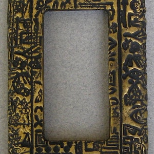 May include: A gold and black decorative wall plate with an intricate design. The plate has a rectangular opening in the center.