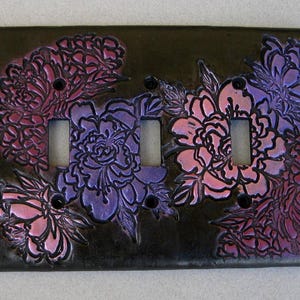 May include: A rectangular triple light switch wall plate featuring a black base with embossed floral patterns in shades of metallic purple and pink. The design includes three vertical rectangular cutouts for switches and four screw holes.