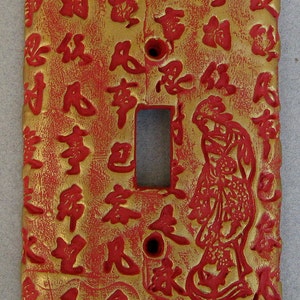 May include: A red and gold light switch cover with a decorative design of Chinese characters and a stylized figure. The cover is made of ceramic and has a textured surface.