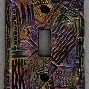 May include: A decorative light switch cover with a textured, abstract design. The cover features a rectangular opening for the switch and two screw holes. The color palette includes shades of purple, gold, and black, with a metallic sheen.