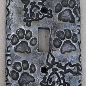 May include: A silver light switch cover with a black paw print design and a black dog silhouette. The cover has a decorative swirl pattern.