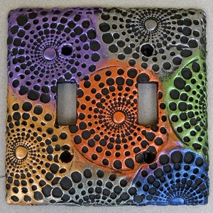 May include: A decorative light switch cover with a geometric pattern in shades of purple, orange, green, and blue. The pattern is made up of concentric circles of dots.
