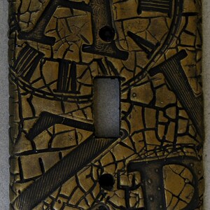May include: Gold light switch cover with a cracked, textured surface and black embossed letters and numbers. The switch cover has a rectangular shape and a single switch opening.