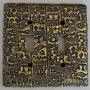 May include: A gold and black light switch cover with a decorative pattern of hieroglyphics. The switch cover has two toggle switches.