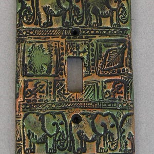 May include: A rectangular light switch plate featuring a textured, multi-colored finish in shades of green, bronze, and black. The design includes embossed elephant figures at the top and bottom, with central panels displaying various botanical and animal motifs.