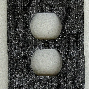 May include: Black outlet cover with a textured surface. The cover has two openings for electrical outlets.