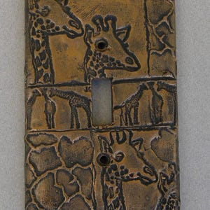 May include: A rectangular bronze-colored light switch plate featuring a textured, embossed design of multiple giraffes and their characteristic spotted patterns. The plate includes a central vertical cutout for a toggle switch and two screw holes.