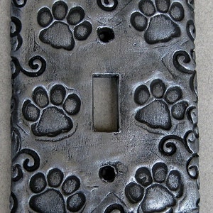 May include: A decorative light switch cover with a silver finish. The cover features embossed paw prints and swirling scrollwork. The switch opening is rectangular, and there are two screw holes. A unique home decor item for pet lovers.