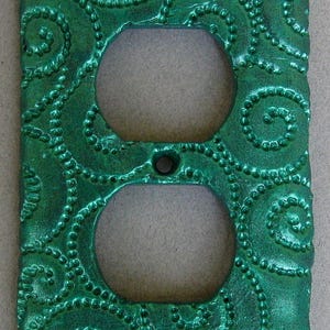 May include: A green outlet cover with a decorative swirl pattern. The cover has a metallic finish and a raised design.