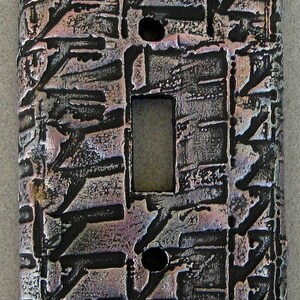 May include: Textured, metallic light switch cover with a unique, abstract design. The cover features a rectangular opening for the switch and two circular holes. The color scheme includes shades of gray, black, and hints of purple and pink.