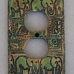 May include: A rectangular electrical outlet wall plate featuring a textured, metallic green and bronze finish. The surface is embossed with detailed illustrations of elephants and small lizard-like creatures, framed by geometric patterns and borders.