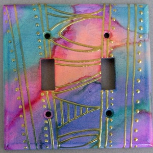 May include: A double light switch cover with a colorful abstract design. The design features pink, purple, blue, and gold accents. The cover is decorated with gold lines and dots.