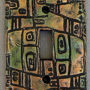 May include: A light switch cover with a geometric pattern in shades of green, brown, and black. The cover has a textured surface and a raised design.