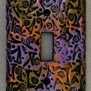 May include: A decorative light switch cover with a textured surface featuring a pattern of swirls and geometric shapes in shades of black, purple, orange, and gold.
