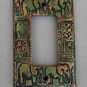 May include: A rectangular light switch cover plate featuring a raised relief design with elephants and various animal motifs. The surface has a metallic finish with blended green, bronze, and black tones, including two screw holes for installation.