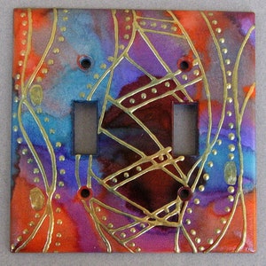 May include: A double light switch cover with a colorful abstract design. The design features a mix of purple, blue, red, and orange colors with gold accents. The gold accents are in the form of lines and dots.