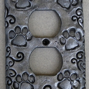 May include: A decorative, silver-colored electrical outlet cover featuring embossed paw prints and swirling patterns. The cover has two circular openings for outlets and a small, central screw hole.