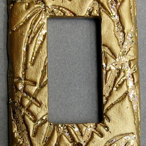 May include: Gold-colored light switch cover with a raised design of bamboo leaves and glitter accents.