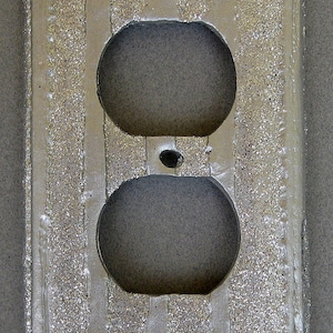 May include: A silver-colored, textured electrical outlet cover with two circular openings for plugs. The cover has a rectangular shape with vertical striped detailing and a small hole between the plug openings.
