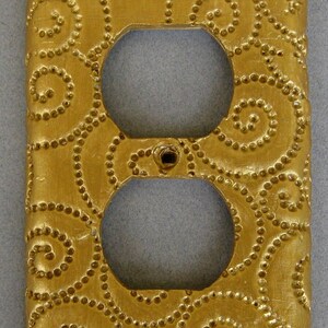 May include: Gold-colored outlet cover with a decorative pattern of swirling circles. The cover is made of a textured material and has a slightly raised design.