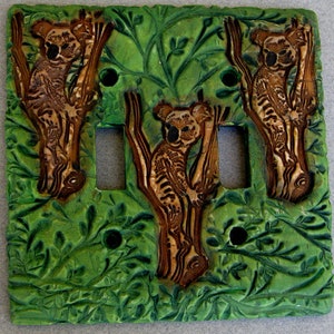 May include: A decorative light switch cover featuring three koalas in trees against a green leafy background. The koalas and trees are brown, with the background in varying shades of green. The cover has three switch holes and three screw holes.