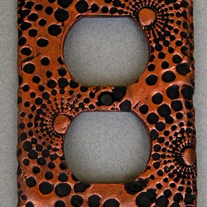 May include: A copper-colored outlet cover with a textured design of black circles and swirls.