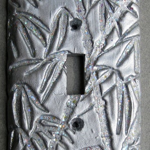 May include: A silver light switch cover with a raised design of bamboo leaves and stems. The cover has a glittery finish.