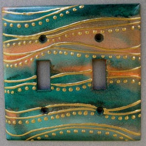 May include: A double light switch cover with a decorative design of wavy lines and gold dots on a teal and peach background.