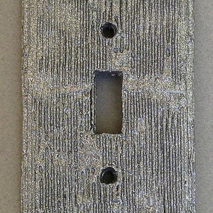 May include: A silver light switch cover with a textured surface that resembles wood grain.