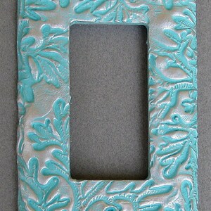May include: A decorative light switch plate with a textured, off-white base and turquoise leaf-like designs. The rectangular plate has a central opening and two screw holes. The design is raised, giving it a 3D effect.