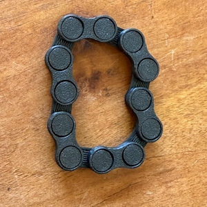 Op de afbeelding: Een zwarte fietsketting in de vorm van de letter "D". De ketting is gemaakt van verbonden cirkelvormige schakels en ligt op een houten oppervlak. De ketting is donkergrijs.