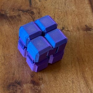 May include: A multi-coloured cube puzzle in shades of blue and purple. The cube is constructed from numerous small, interconnected blocks, forming a distinctive geometric form. The puzzle is designed for manipulation and reconfiguration.