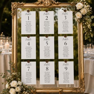May include: A large, ornate gold-toned picture frame displaying nine white rectangular seating chart cards arranged in a grid. Each card features the text table followed by a number and a list of names, secured with small brown wax seals.