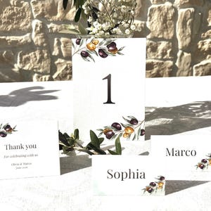 Puede incluir: Decoración de mesa de boda con tarjetas blancas con ilustraciones de ramas de olivo. Incluye una tarjeta con el número 1, tarjetas con nombres y una tarjeta de agradecimiento. Las tarjetas están decoradas con ramas de olivo en acuarela.