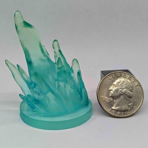 Puede incluir: Una escultura translúcida de color turquesa que se asemeja a una explosión de energía o agua. La escultura está montada sobre una base circular. Hay una moneda al lado para comparar el tamaño.