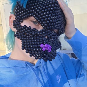 May include: A black beaded mask with a purple accent on the lower part of the face. The mask covers the eyes and nose. The person is wearing a blue hoodie with a white Nike logo.