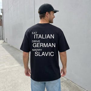 May include: Black t-shirt with white text: "EAT ITALIAN DRIVE GERMAN MARRY SLAVIC." The shirt is worn with a black cap and blue jeans. The person is standing in front of a gray wall.