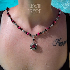 Può includere: Una collana con un ciondolo centrale dal design a maglia metallica argentata e una singola perla rosa, collegata a una catena di perle rosa, perle nere e maglie metalliche argentate alternate. Il testo ELEVENTH RAVEN è visibile.