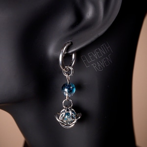 Puede incluir: Un pendiente plateado con un aro, una cuenta azul y transparente, y una estructura plateada en forma de jaula que sostiene otra cuenta azul y transparente. El texto "ELEVENTH RAVEN" es visible.