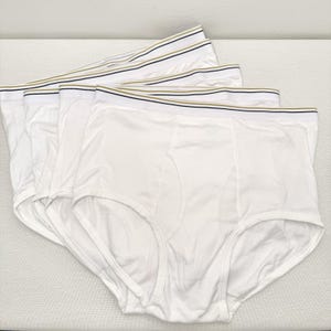 May include: A stack of white briefs featuring a wide elastic waistband. The waistband is white with thin horizontal stripes in black and gold. Each pair of underwear is constructed with a classic fly front design and finished with white stitching.