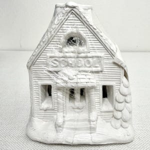 May include: A white ceramic schoolhouse figurine featuring a gabled roof, a central entrance with a small porch, and a sign above the door with the word SCHOOL embossed in capital letters. The structure is wrapped in clear plastic film.