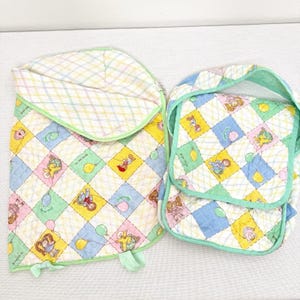 May include: Two-piece baby bedding set featuring a quilted design with colorful squares. The squares display cartoon characters, balloons, and other playful elements. The set includes a blanket and a matching bib, both trimmed in green.