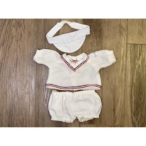 May include: A white doll outfit consisting of a short-sleeved shirt, bloomers, and a visor. The shirt features a V-neckline with red and blue trim, and the hem and sleeves are finished with matching red and blue stripes. The visor is plain white.