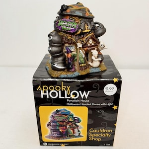 May include: A Spooky Hollow porcelain house titled Cauldron Specialty Shop, featuring a witch, ghosts, and stacked black cauldrons. The structure has a purple sign with green text, a dark roof, and sits atop its original black cardboard box.