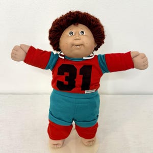 May include: A doll with brown yarn hair and a round face featuring dimples. The doll wears a red and teal long-sleeved shirt with the number 31 printed in black on the front, paired with matching teal and red pants. A white collar is visible.