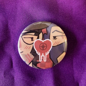 Peut inclure: Un badge rond illustré représentant deux personnages aux joues roses. Au centre, un motif de cœur rouge avec un symbole de cible en forme de croix noire et un effet de goutte est affiché de manière proéminente sur les deux personnages.